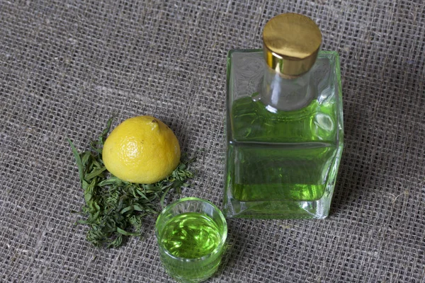 Silna Nalewka na Tarhuna w przezroczystej prostokątnej butelce. Pić jasny zielony wlewa się do szklanki. Obok składników do gotowania. Liści Estrada i cytryna. — Zdjęcie stockowe