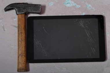Kırık bir ekrana sahip tablet, yüzeyde eski püsyen bir boya ile yer alır. Yakınlarda bir çekiç var..