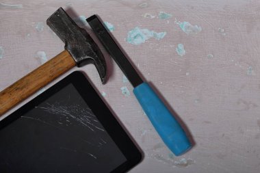 Kırık bir ekrana sahip tablet, yüzeyde eski püsyen bir boya ile yer alır. Yakındaki bir çekiç ve keski.
