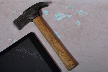Kırık bir ekrana sahip tablet, yüzeyde eski püsyen bir boya ile yer alır. Yakınlarda bir çekiç var..