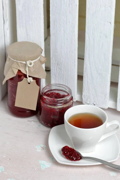 Jars of jam, covered with paper and tied with twine. On the string hang craft labels. One jar is open. Near a cup of tea. — Stock Photo, Image