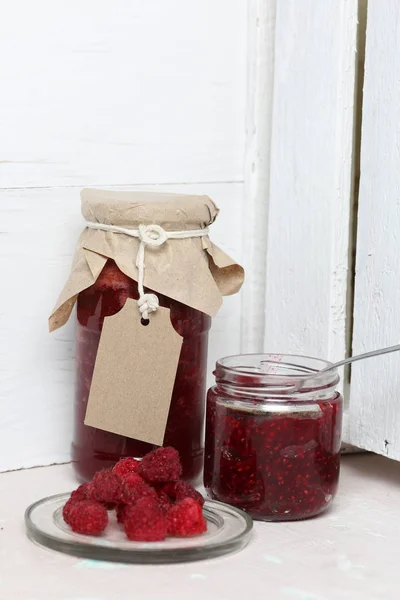 Jars of jam, covered with paper and tied with twine. On the string hang craft labels. One jar is open. Next to a plate of fresh raspberries.