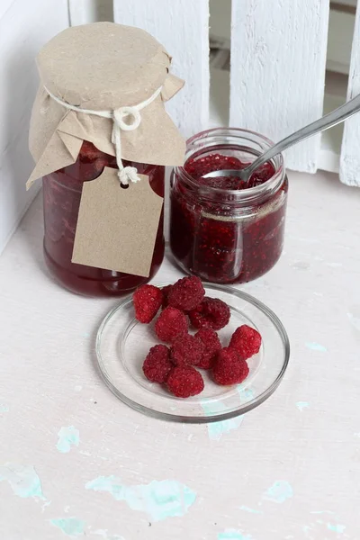 Jars of jam, covered with paper and tied with twine. On the string hang craft labels. One jar is open. Next to a plate of fresh raspberries.