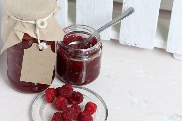 Jars of jam, covered with paper and tied with twine. On the string hang craft labels. One jar is open. Next to a plate of fresh raspberries.