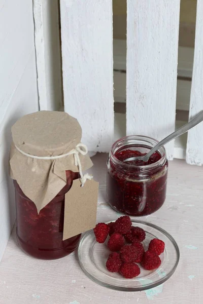 Jars of jam, covered with paper and tied with twine. On the string hang craft labels. One jar is open. Next to a plate of fresh raspberries.