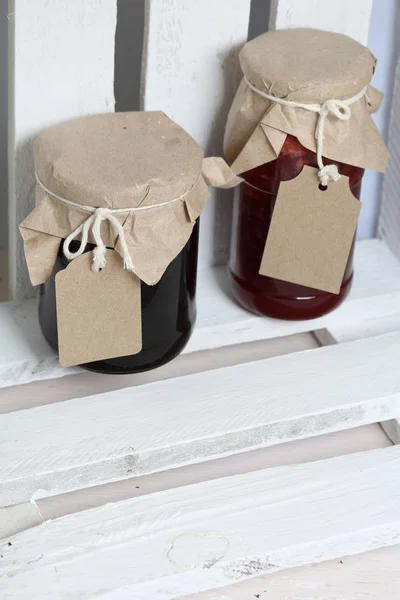 Frascos de mermelada, cubiertos de papel y atados con cordel. En la cuerda cuelgan etiquetas artesanales. Stand en una caja de madera . — Foto de Stock