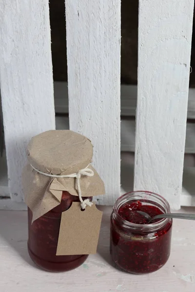 Frascos de mermelada, cubiertos de papel y atados con cordel. En la cuerda cuelgan etiquetas artesanales. Un frasco está abierto . — Foto de Stock