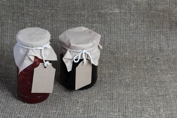 Vasetti di marmellata, ricoperti di carta e legati con spago. Sulla corda appendere etichette artigianali. Sullo sfondo di lino tessuto grossolano . — Foto Stock