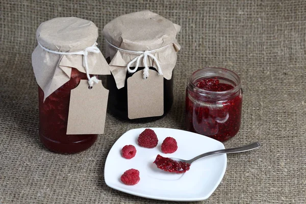 Jars of jam, covered with paper and tied with twine. On the string hang craft labels. One jar is open. Near fresh raspberries on a plate.