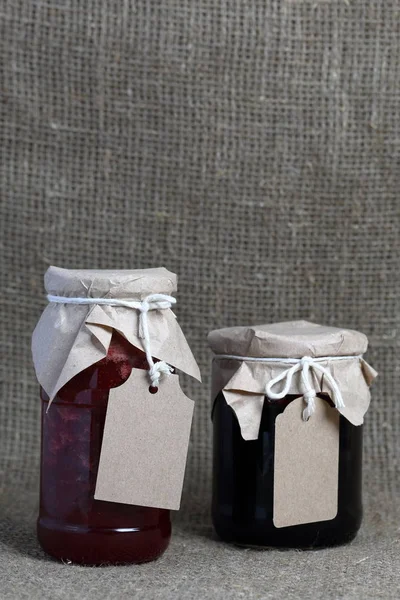 Pots de confiture, recouverts de papier et attachés avec de la ficelle. Sur la ficelle accrocher des étiquettes artisanales. Sur fond de toile de lin tissu grossier . — Photo