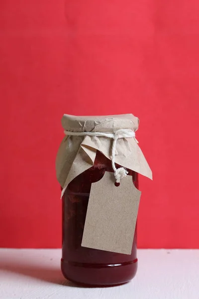 Jars of jam, covered with paper and tied with twine. On the string hang craft labels. Against the background of coral color. — Stock Photo, Image