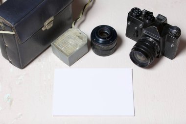 Eski film kamera, çıkarılabilir lens ve kablolu flaş. Masanın yüzeyinde yatıyorlar. Beyaz bir fotoğraf kağıdının yanında.