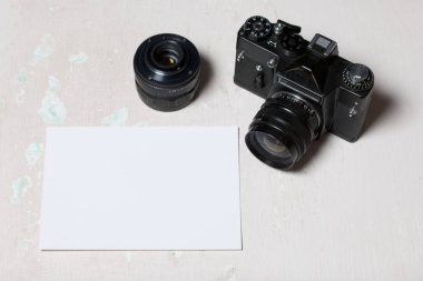 Eski film kamera ve çıkarılabilir lens. Masanın yüzeyinde yatıyorlar. Beyaz bir fotoğraf kağıdının yanında.