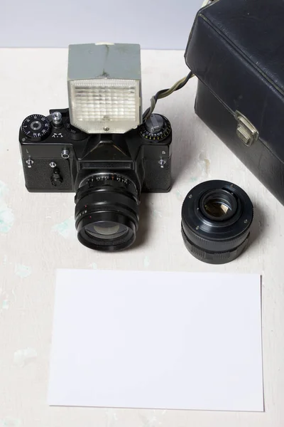 Old film camera, removable lens and wired flash. They lie on the surface of the table. Near a white sheet of photo paper.