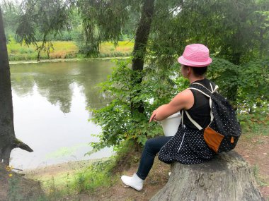 Kafasında şapka ve omuzlarında sırt çantası olan kadın mantar toplayıcısı. Bir orman evinde bir kütüğün üzerine oturdu ve nehre baktı..