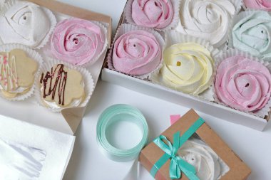 Marshmallows farklı renk ve şekillerde. Bazıları çiçek şeklinde. Hediye kutularına yığılmış. Yakınlarda pansuman kutuları için renkli kurdeleler bulunmaktadır. Yukarıdan görüntüle.