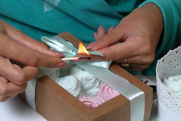 Woman puts marshmallows of different colors into gift boxes. Bandages boxes with a colored ribbon. Tie a ribbon on a bow and cauterize the edge with a burning match.