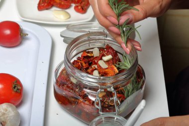 Bir kavanozda baharat ve sarımsak ile güneşte kurutulmuş domates. Kadın biberiye ekler. Biberiye, domates ve sarımsak yakınında.