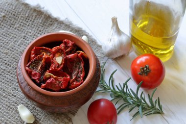 Bir kil tencerede baharat ve sarımsak ile güneşte kurutulmuş domates. Yakınlarda zeytinyağı, domates, biberiye ve sarımsak içeren bir şişe var..