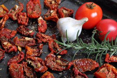 Bir fırın tepsisine baharat ve sarımsak ile güneşte kurutulmuş domates. Yakınlarda taze biberiye, domates ve bir baş sarımsak var..