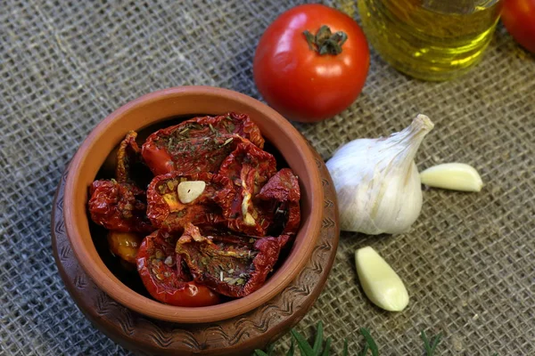 Sun-dried tomatoes with spices and garlic in a clay pot. Nearby is a bottle with olive oil, tomatoes, rosemary and garlic. — Stock Photo, Image