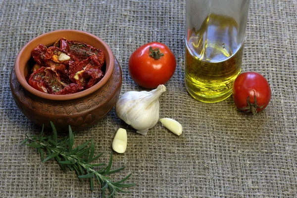 Suszone pomidory z przyprawami i czosnkiem w glinianych garnku. W pobliżu znajduje się butelka z oliwą z oliwek, pomidory, rozmaryn i czosnek. — Zdjęcie stockowe