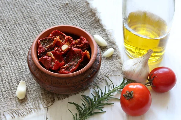 Tomates secados pelo sol com temperos e alho em um pote de barro. Perto está uma garrafa com azeite, tomate, alecrim e alho . — Fotografia de Stock