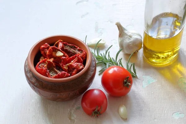 Tomates secados pelo sol com temperos e alho em um pote de barro. Perto está uma garrafa com azeite, tomate, alecrim e alho . — Fotografia de Stock