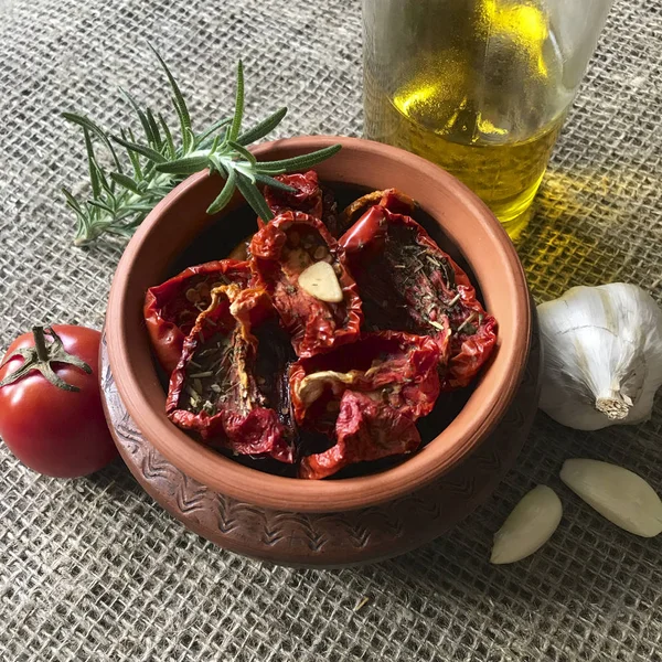 Sun-dried tomatoes with spices and garlic in a clay pot. Nearby is a bottle with olive oil, tomatoes, rosemary and garlic. — Stock Photo, Image