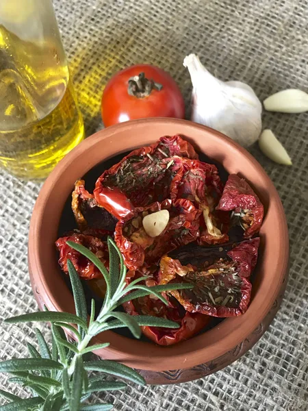 Sun-dried tomatoes with spices and garlic in a clay pot. Nearby is a bottle with olive oil, tomatoes, rosemary and garlic. — Stock Photo, Image