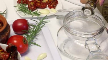 Baharat ve sarımsak ile güneşte kurutulmuş domates. Onları korumak için, bir kadın bir kavanoza zeytinyağı döker, tuz ve baharat ekler. Yakınlarda taze biberiye, domates ve sarımsak var..