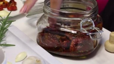 Bir kadın kavanoza kurutulmuş domates ekler. Biberiye ve baharat dalvardır. Yakınlarda taze biberiye, domates ve bir baş sarımsak var..