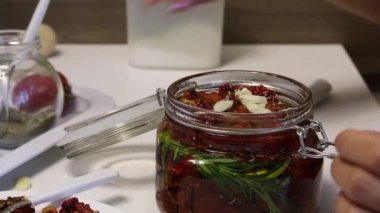 Bir kadın kavanoza sarımsak ve kurutulmuş domates ekler. Biberiye, sarımsak ve baharat dalvardır. Zeytinyağı ile dökülür.