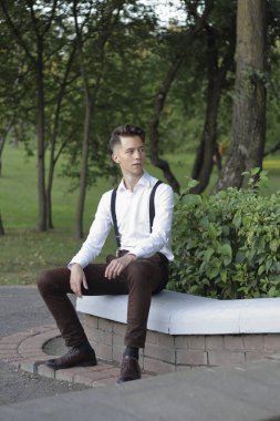 Şık giyimli genç adam bir parkta poz. Kaldırıma oturur ve uzağa bakar..