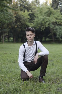 Şık giyimli genç adam bir parkta poz. Çimlere oturur ve uzağa bakar..