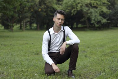 Şık giyimli genç adam bir parkta poz. Çim oturur ve bir gülümseme ile kameraya bakar.