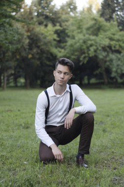 Şık giyimli genç adam bir parkta poz. Çimlere oturur ve uzağa bakar..