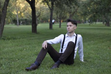 Şık giyimli genç adam bir parkta poz. Çimlere oturur ve uzağa bakar..