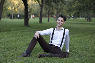 Şık giyimli genç adam bir parkta poz. Çim oturur ve bir gülümseme ile uzağa bakar.
