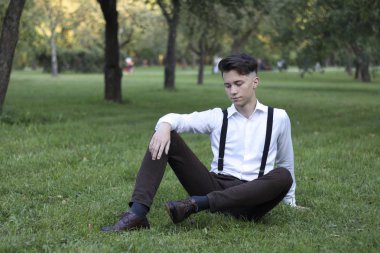 Şık giyimli genç adam bir parkta poz. Çimlere oturur ve uzağa bakar..