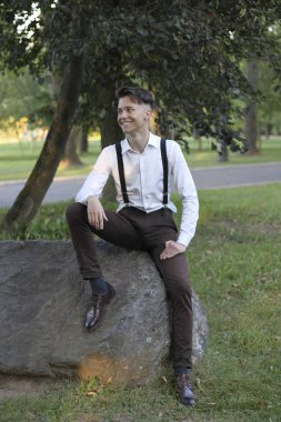 Şık giyimli genç adam parkta bir taş kaya üzerinde oturur. Gülümseyip uzaklara bakıyor..