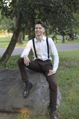 Şık giyimli genç adam parkta bir taş kaya üzerinde oturur. Düşünceli bir bakışla.