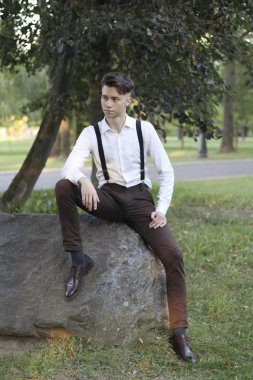 Şık giyimli genç adam parkta bir taş kaya üzerinde oturur. Düşünceli bir bakışla.