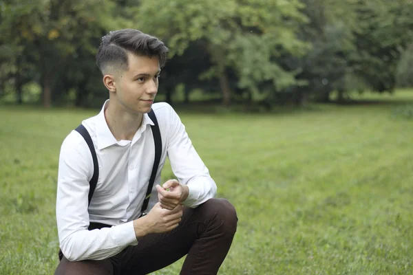 Stijlvol gekleed jonge man poseren in een park. Squatting op het gazon rechtzetten van de knop op zijn shirt. — Stockfoto