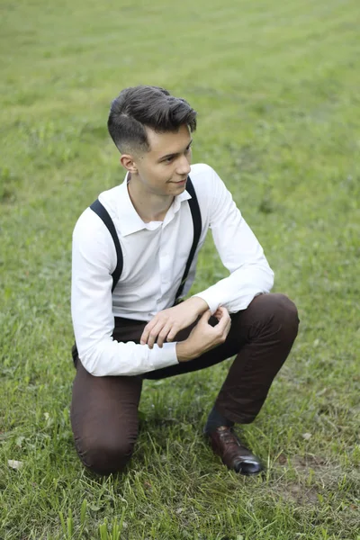 Şık giyimli genç adam bir parkta poz. Çimlere çömelmek gömleğindeki düğmeyi düzeltir..