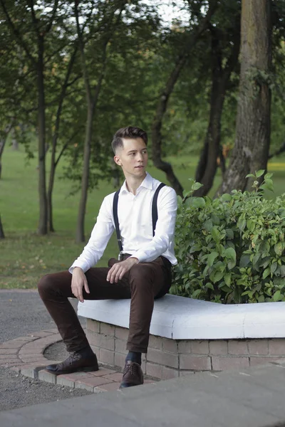 Şık giyimli genç adam bir parkta poz. Kaldırıma oturur ve uzağa bakar..
