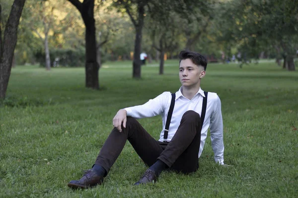 Şık giyimli genç adam bir parkta poz. Çimlere oturur ve uzağa bakar..