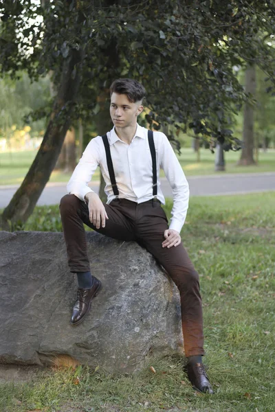 Şık giyimli genç adam parkta bir taş kaya üzerinde oturur. Düşünceli bir bakışla.