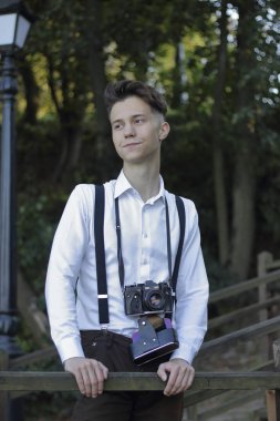 Şık giyimli genç adam bir parkta poz. Bir Slr film kamera boyun asılı.