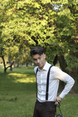 Şık giyimli genç adam bir parkta poz. Kameraya şaşkınlıkla bakıyor, pantolonunu düzeltir..